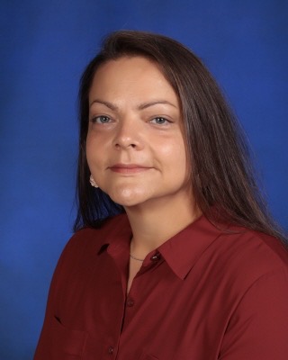 A woman with long dark hair and a serious expression is wearing a maroon blouse against a blue background.