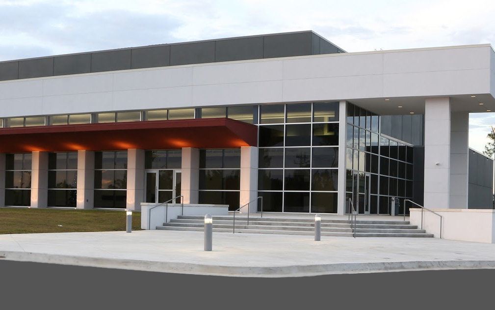 A modern building featuring large glass windows, a prominent entrance with steps, and a sleek design, set against a cloudy sky.