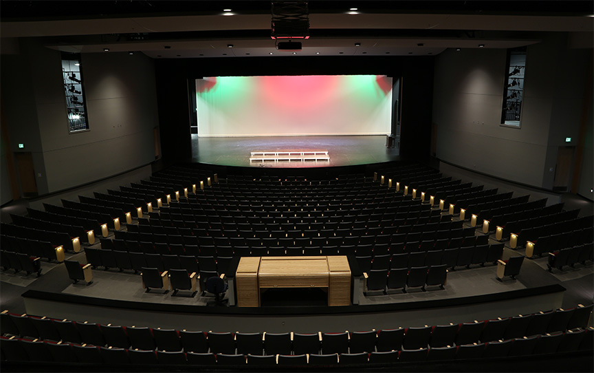 An interior view of a theater featuring rows of empty seats facing a brightly lit stage with colorful lighting.