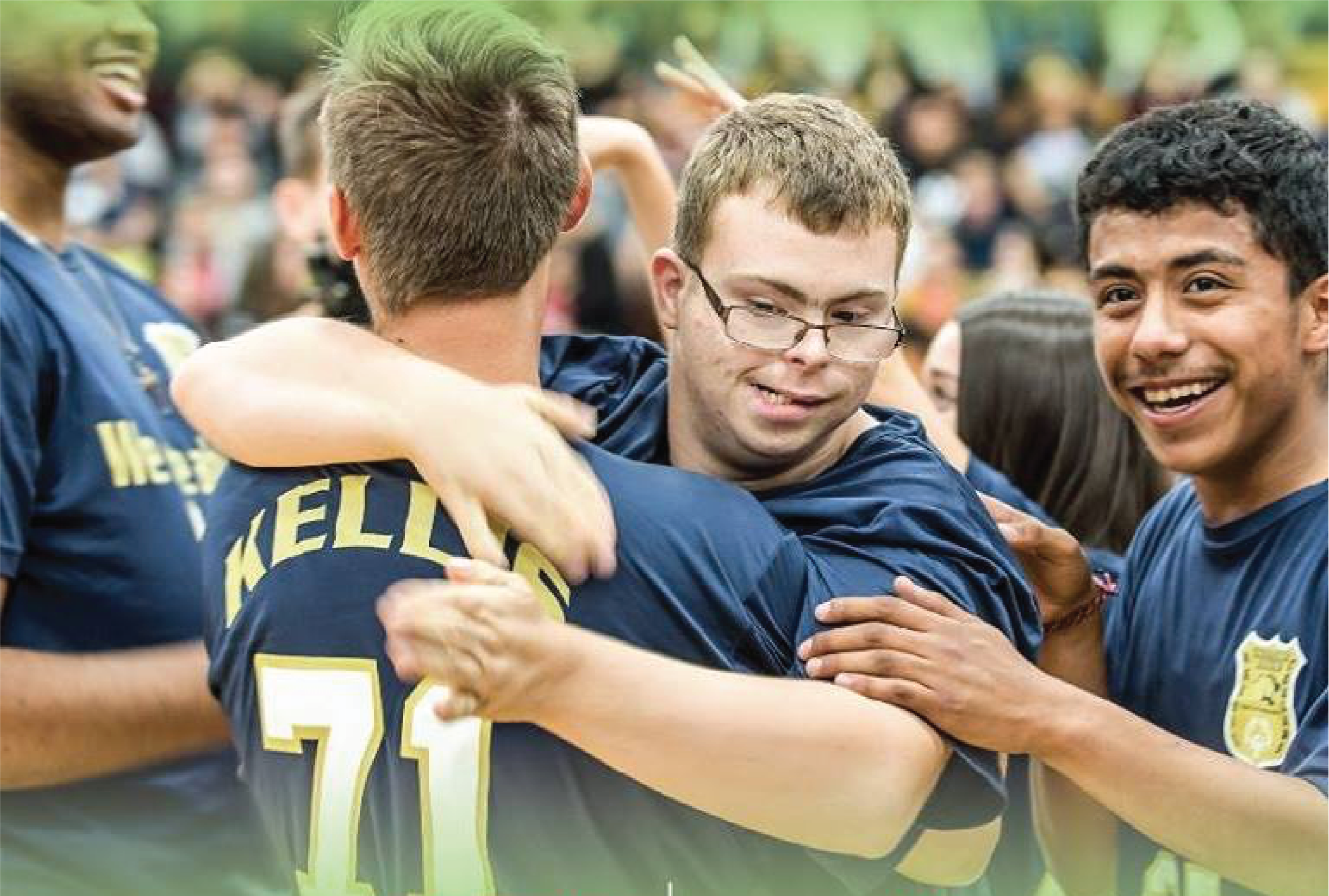 Group of people in matching blue shirts with yellow text, embracing. The central figure's shirt reads 'KELLIS 71'. Some faces are blurred.