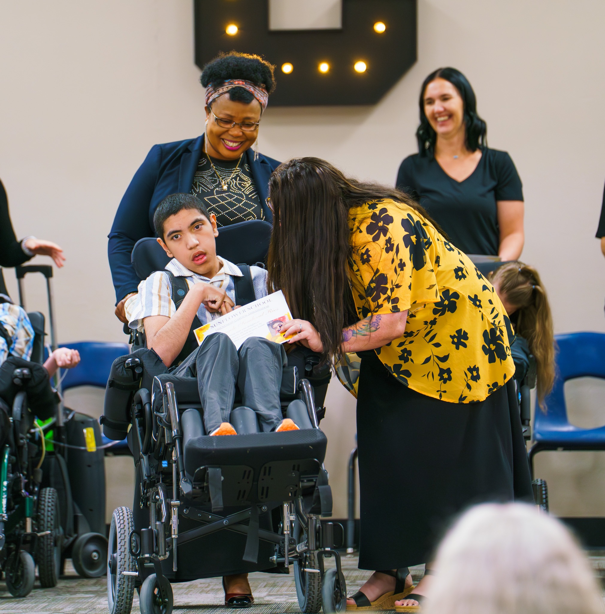 "A group of people gathered together, including individuals in wheelchairs. One person wearing a yellow floral shirt is leaning over to interact with a person in a wheelchair who is holding a certificate."