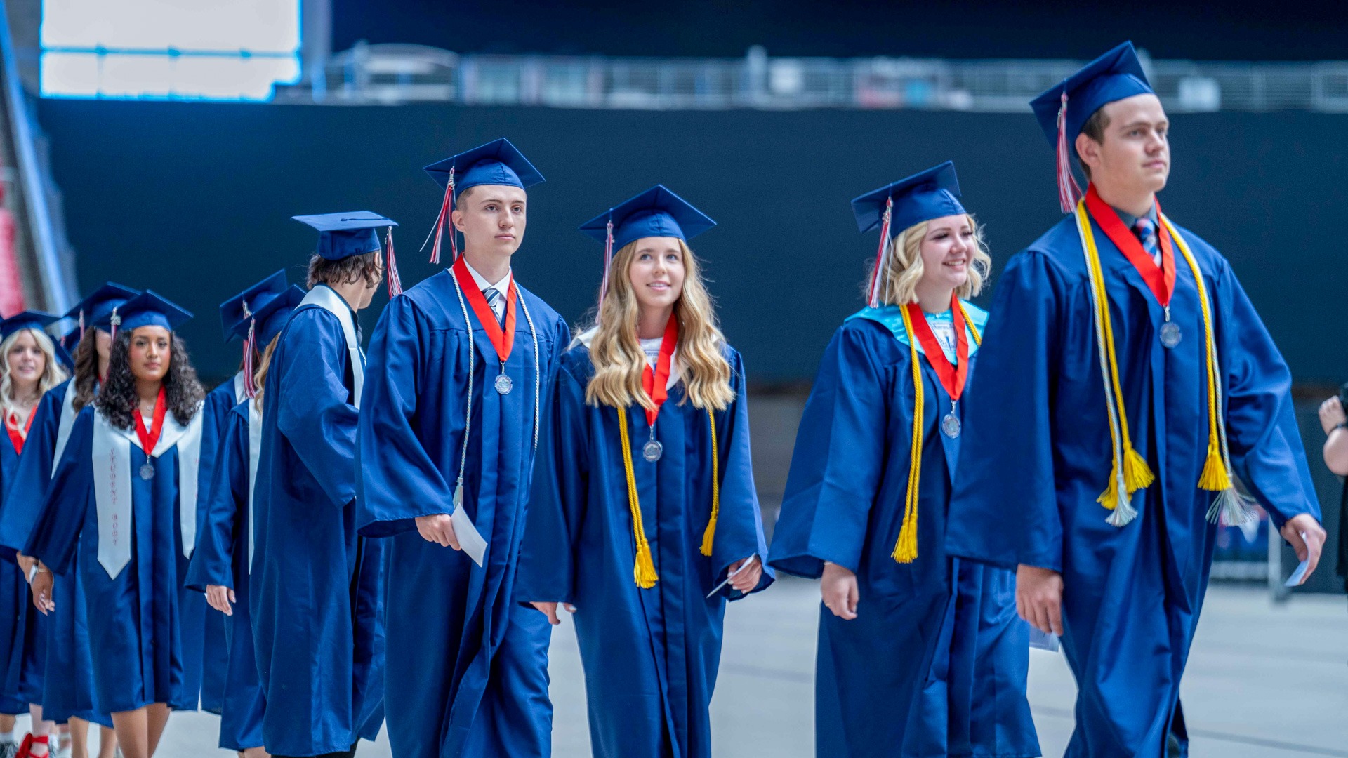 Graduates walking in a blue cap and gown, with red ribbon medallions and yellow cords.