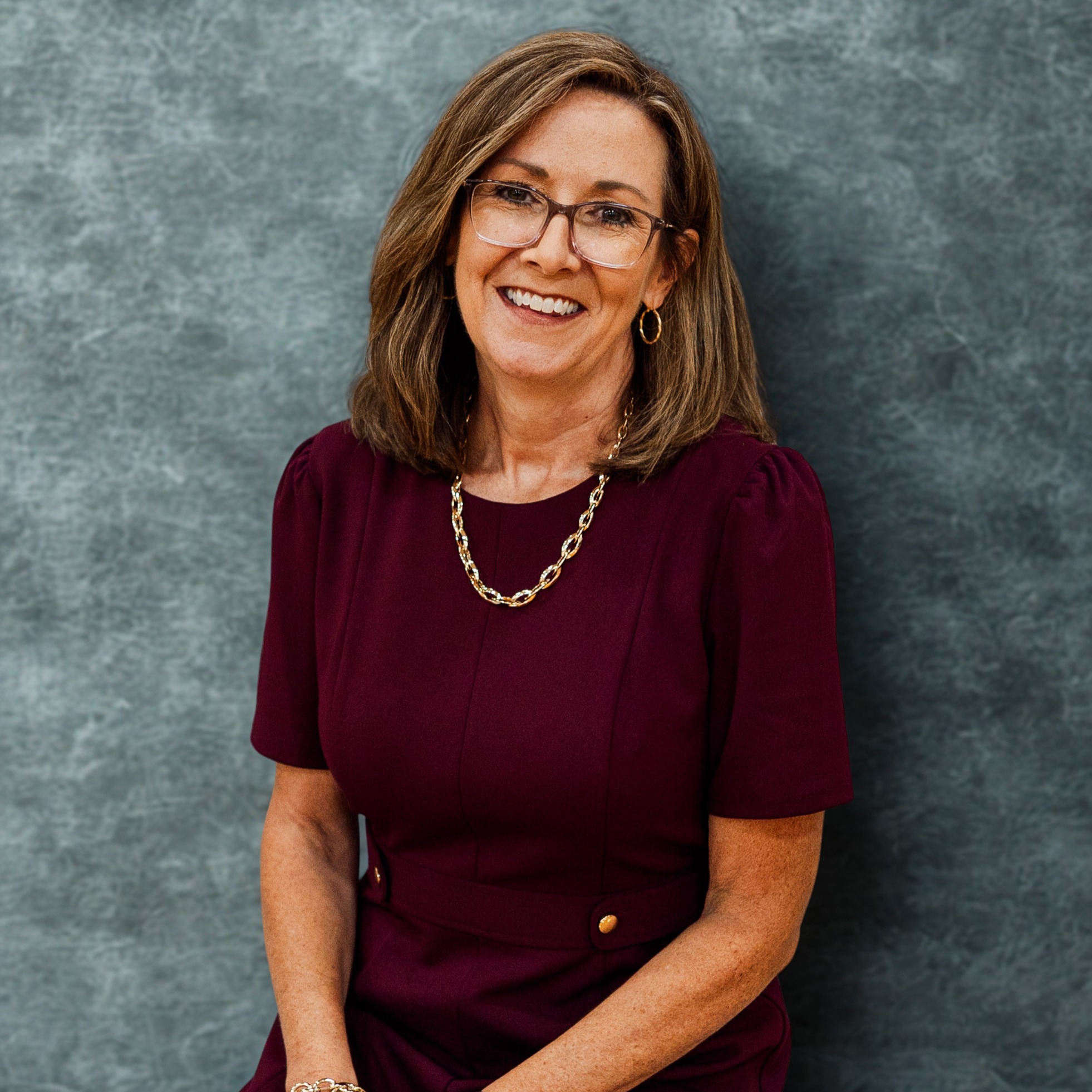 A woman in a maroon dress and glasses sits against a gray wall, smiling. She has a necklace, bracelets, and a watch.