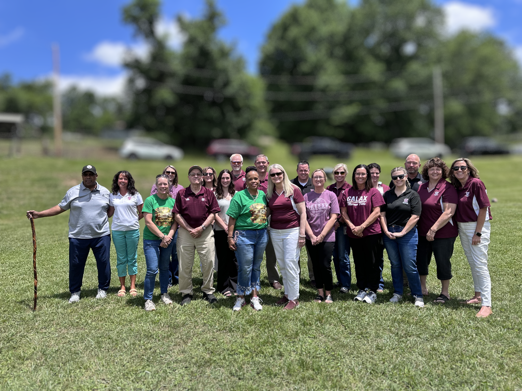 Group of people in matching purple and green shirts and white pants standing in a grassy field.