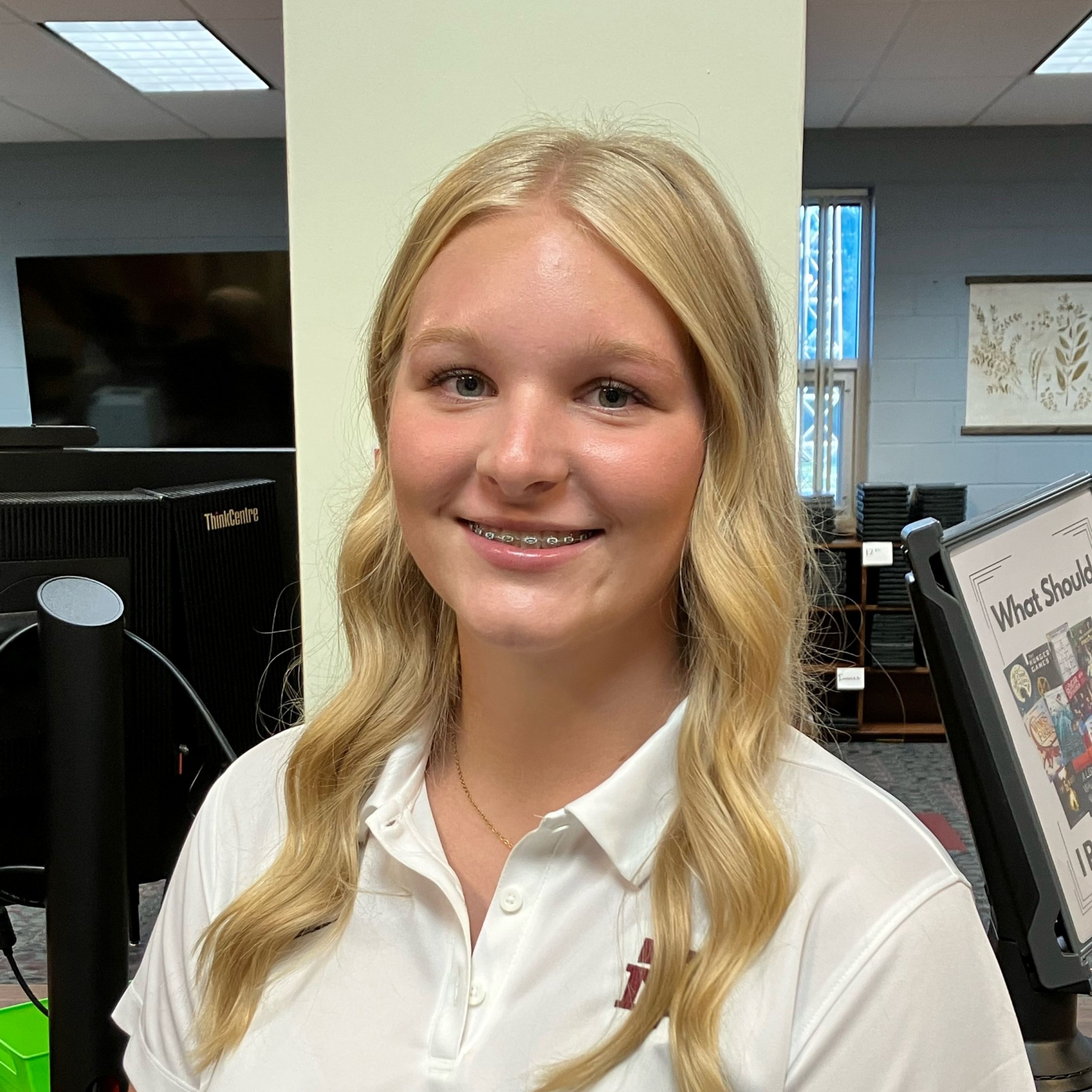 Blonde woman in a white polo shirt stands in front of a computer monitor.