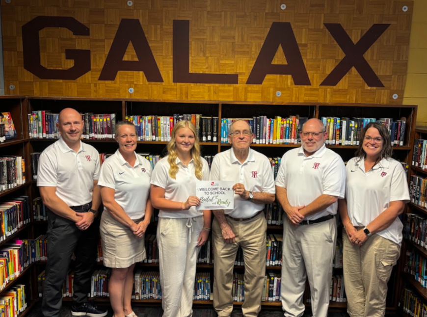Six adults, some in white shirts and khaki pants, pose for a photo in a library.