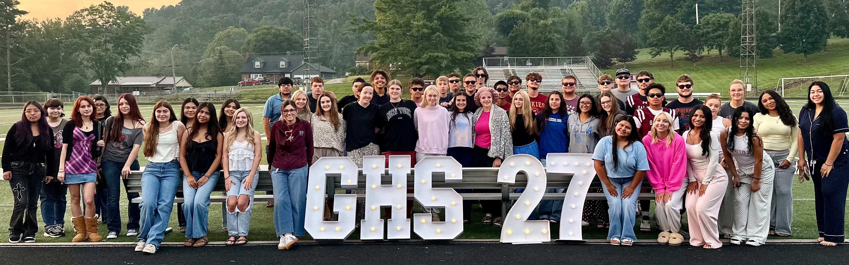 A group of students stands on a track with the letters "GHS 27" in the foreground. Behind them is a field and trees.