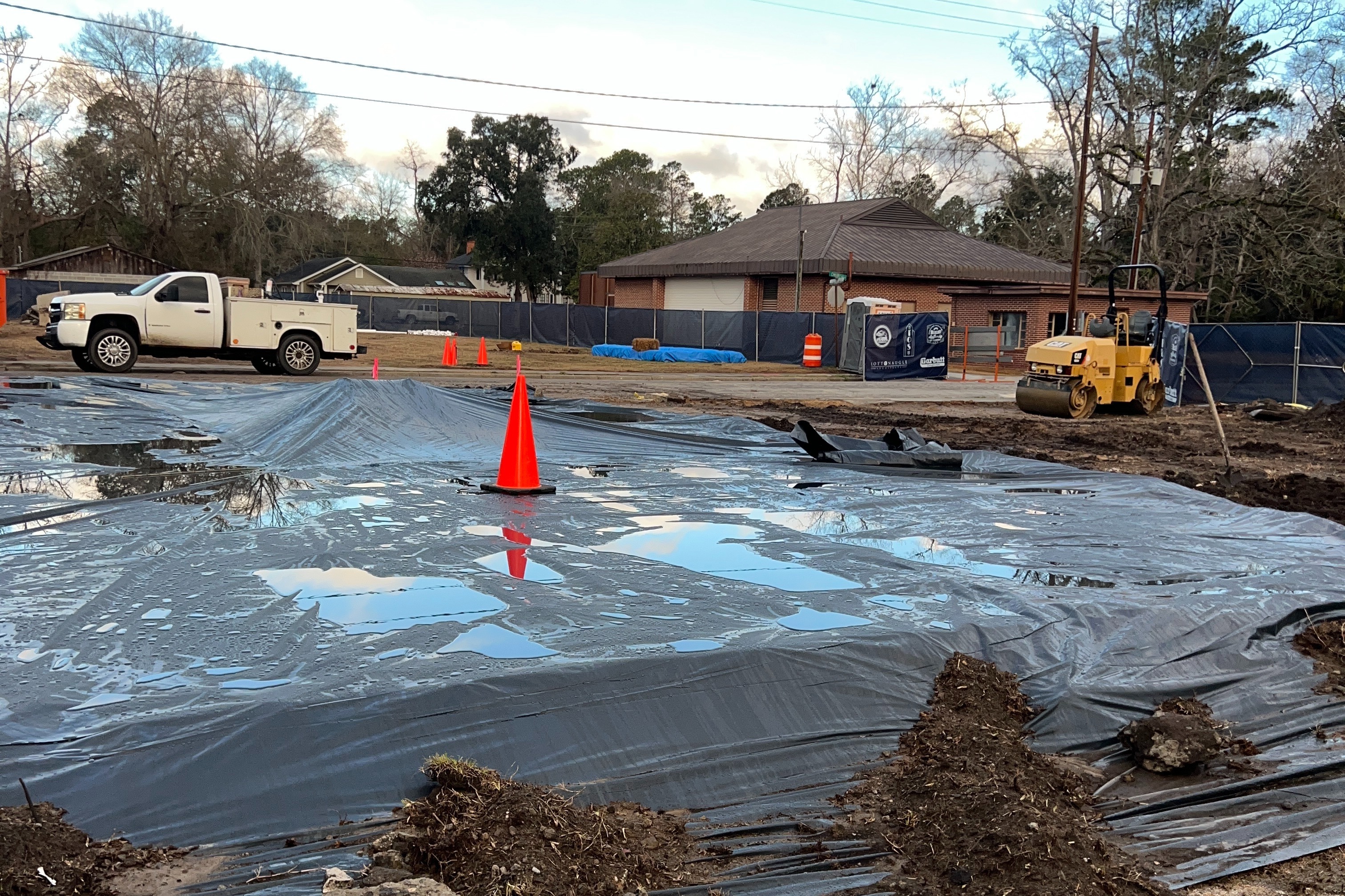 The exposed soil behind the construction site tarped over with a plastic cover held down with small mounds of dirt around the perimeter to protect the soil below from the elements. Puddles on tarp allude to it having recently rained.
