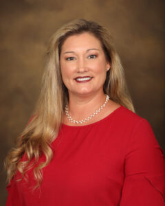 A woman with long, wavy blonde hair wearing a red blouse and a pearl necklace smiles against a blurred brown background.