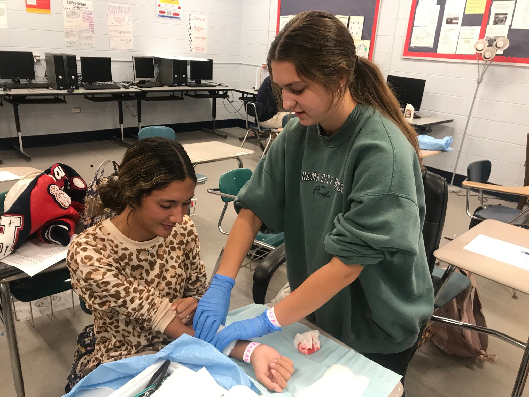 A student in a green sweatshirt is applying a bandage to another student's arm in a classroom setting, with computers visible in the background.