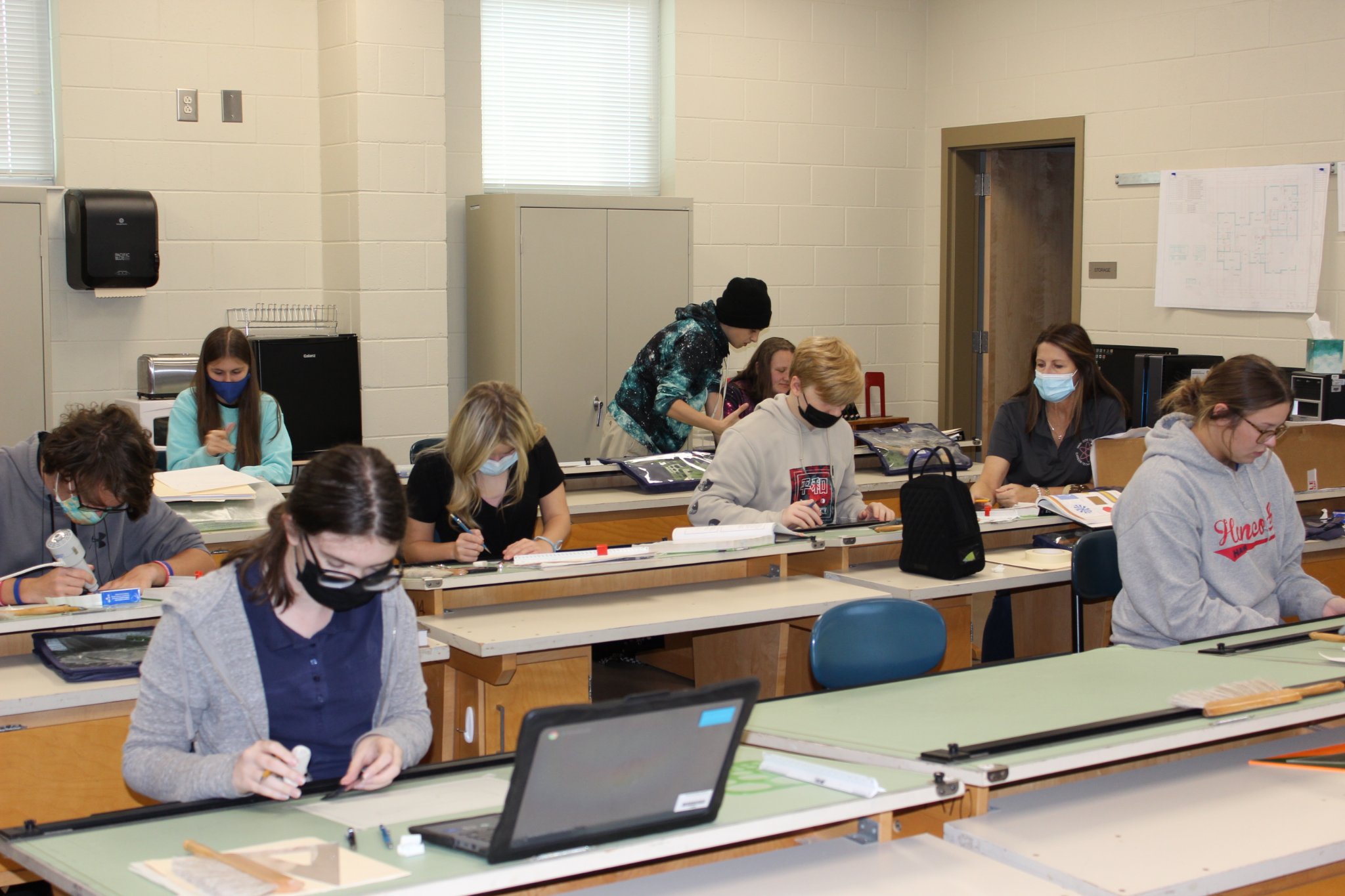 A classroom scene with students working at desks, some wearing masks, engaged in various activities, while a teacher assists in the background.