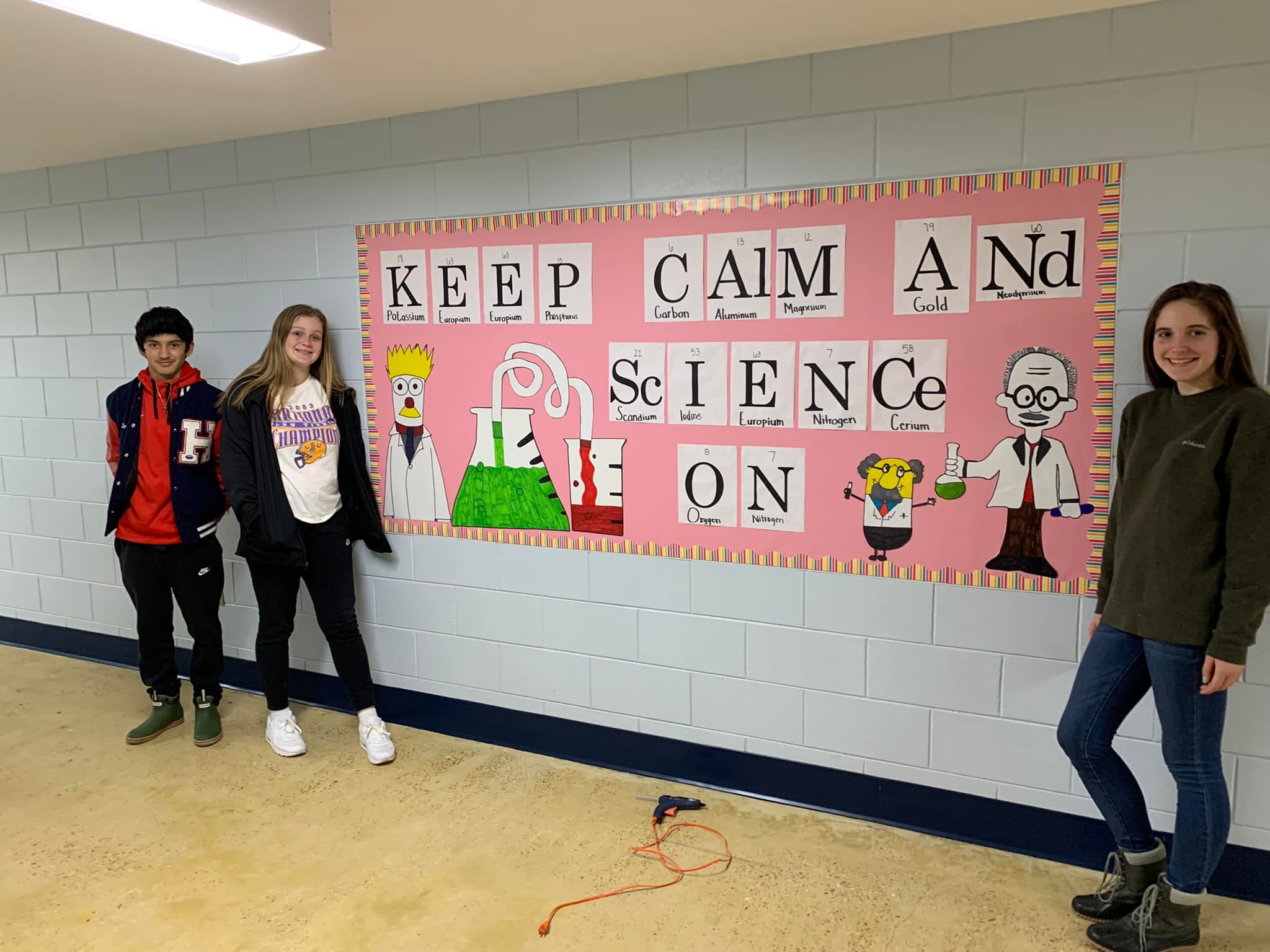 Three students stand in front of a colorful bulletin board that reads 'KEEP CALM AND SCIENCE ON,' featuring cartoon characters related to science.