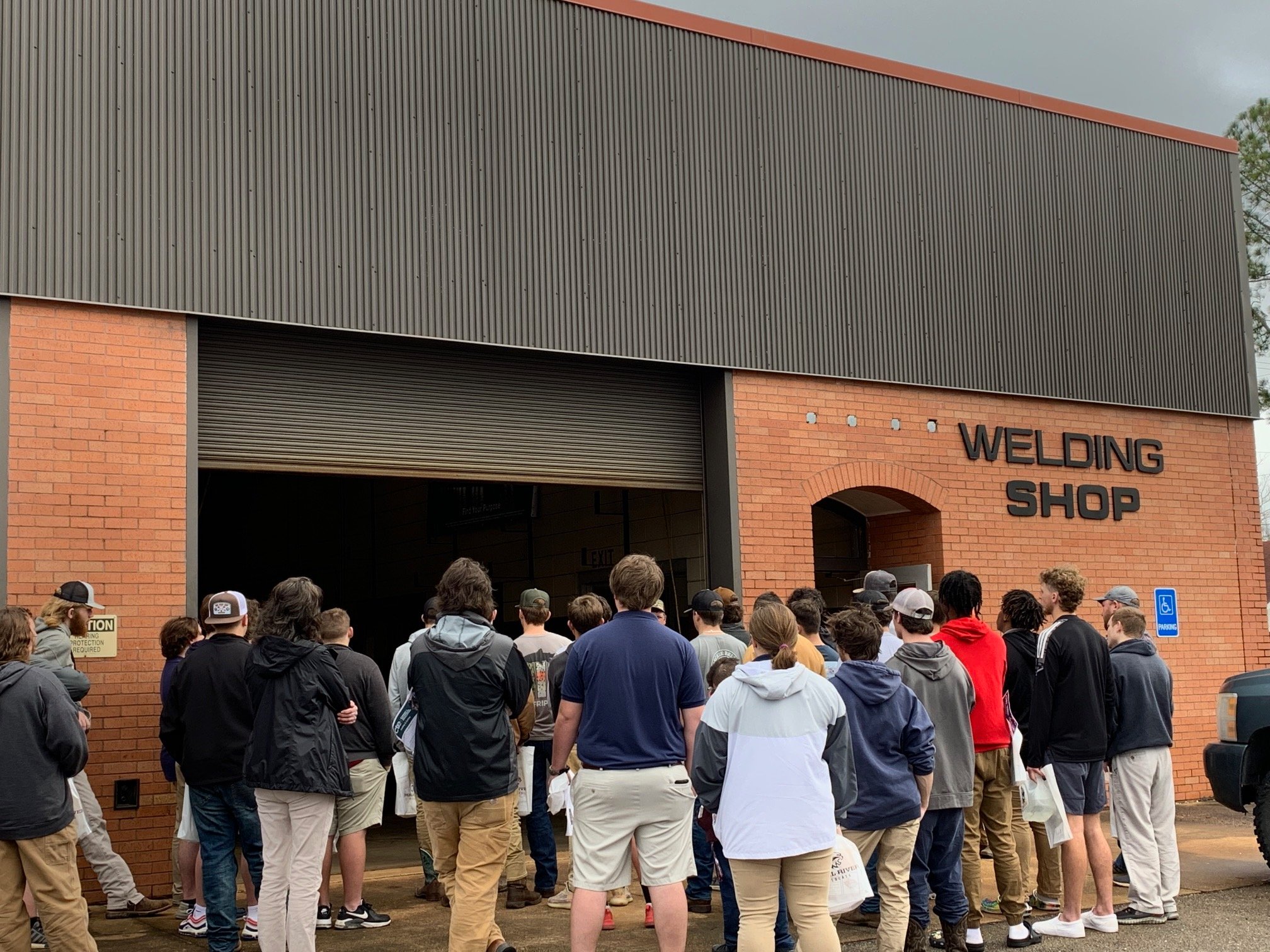 A group of students gathered outside a welding shop building, waiting to enter, with a cloudy sky overhead.