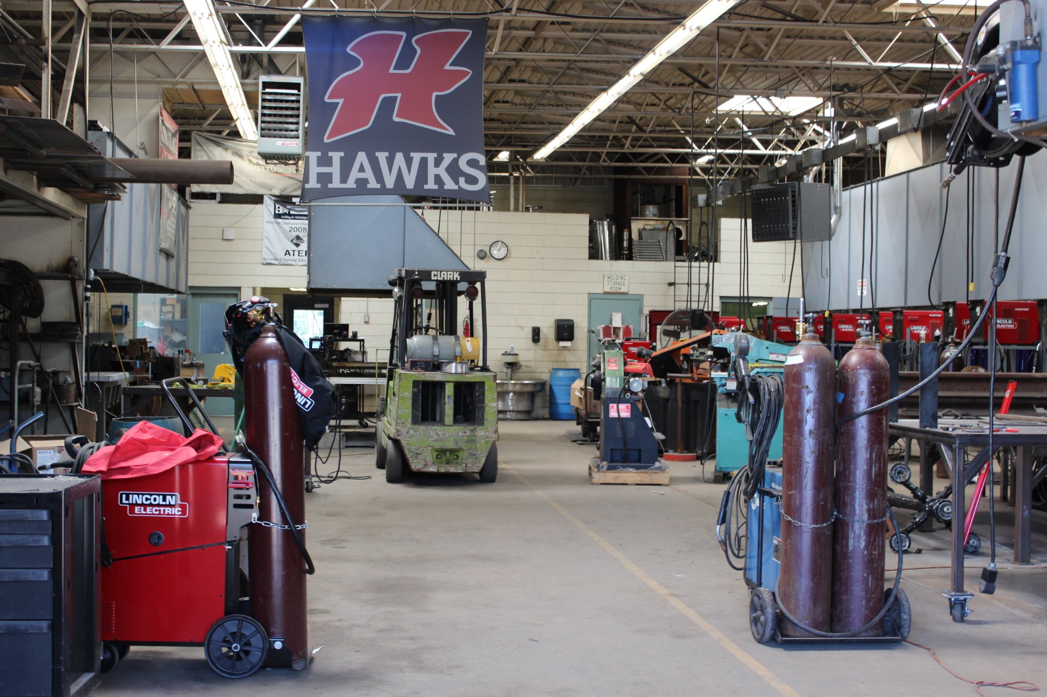 A spacious workshop featuring various equipment, including welding machines and gas cylinders, with a large 'HAWKS' banner hanging from the ceiling.