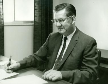 A man in a suit and glasses is seated at a desk, writing on a piece of paper with a pen, in a well-lit office setting.