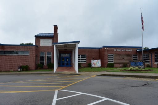 1. A school building's facade featuring a flag atop its roof, symbolizing education and community pride. 2. The front view of a school building, prominently displaying a flag on its roof, representing national pride and learning. 3. An exterior shot of a school building with a flag flying above, highlighting the institution's commitment to education.