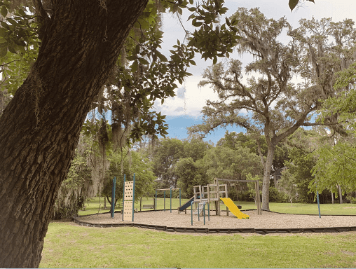 A playground with a slide and climbing structure is surrounded by trees and grassy areas, under a partly cloudy sky.