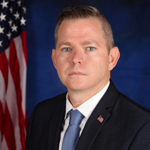 A formal portrait of a man in a suit with a blue tie, standing in front of an American flag and a blue backdrop.