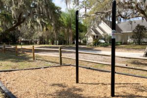 A set of black pull-up bars stands on a bed of wood chips in a park, with a grassy area and houses visible in the background.