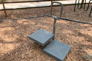 A gray outdoor exercise machine featuring two textured foot platforms and a vertical handle, situated on a bed of wood chips in a park setting.