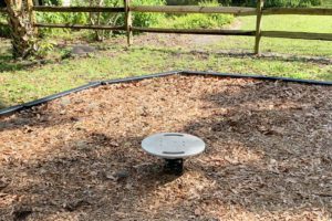 A circular metal platform sits on a bed of wood chips in a grassy area, surrounded by a wooden fence and trees in the background.