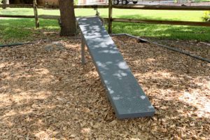 A gray ramp situated in a playground area, surrounded by wood chips and grass, with a tree in the background.