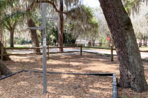 A park area featuring a metal exercise station surrounded by trees and mulch, with a fence in the background.