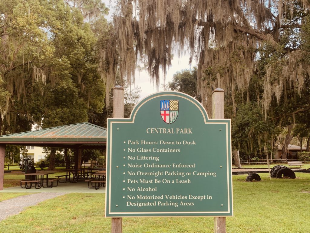 A green sign at Central Park listing park rules and regulations, including restrictions on fishing, camping, and pet leash requirements, with a pavilion visible in the background.