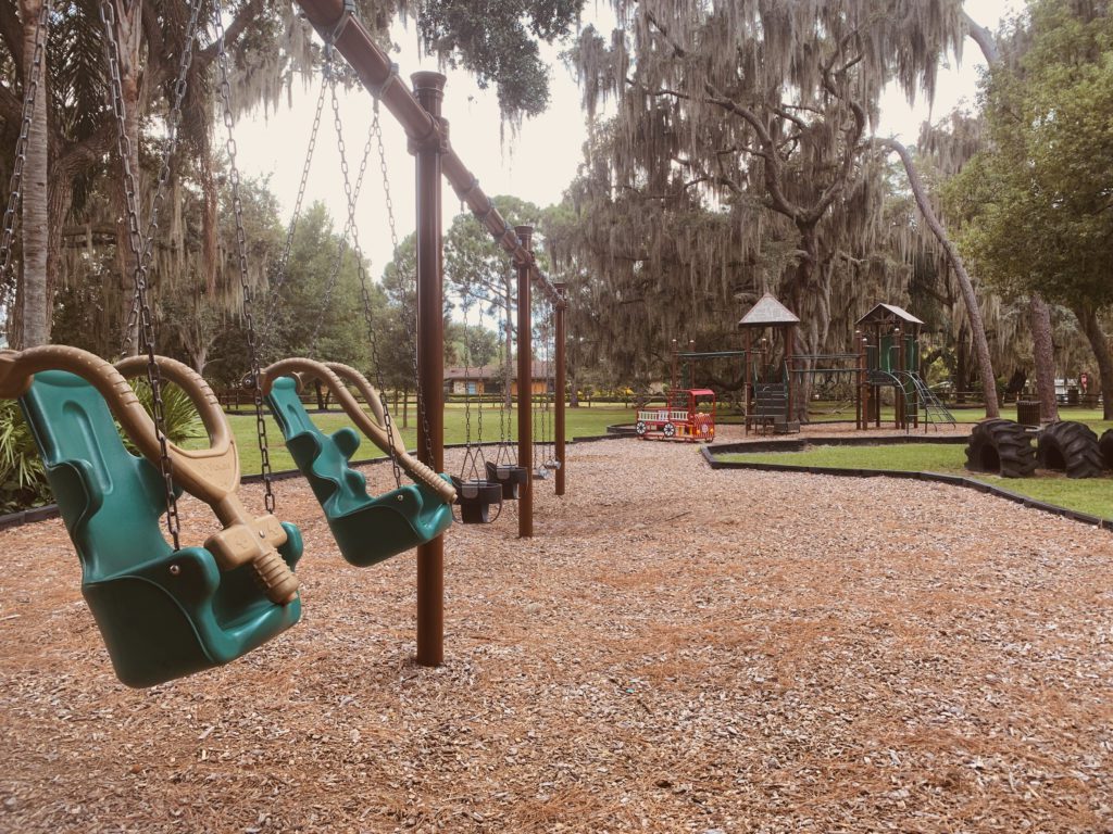 A playground featuring swings with green seats, surrounded by gravel and trees, with additional play structures visible in the background.