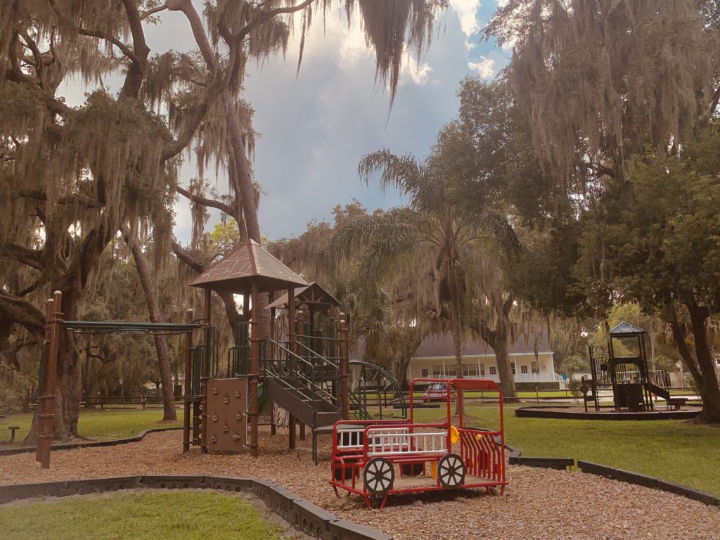 A playground featuring a wooden play structure and a colorful train-themed play area, surrounded by trees with Spanish moss.