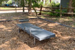 A metal bench sits on a bed of leaves in a park-like setting, surrounded by trees and a wooden fence.