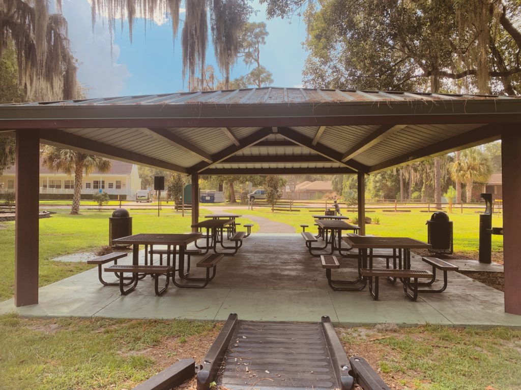 A covered picnic area with multiple tables and trash bins, surrounded by green grass and trees in a park setting.