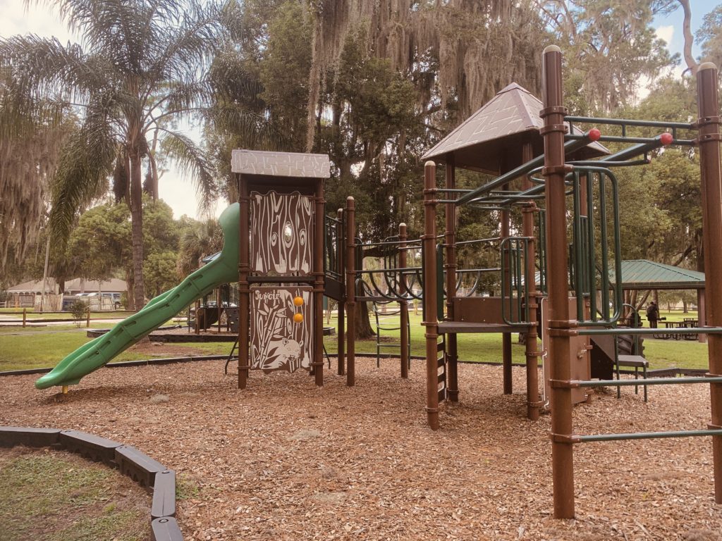 A playground featuring a green slide, climbing structures, and surrounded by a grassy area with trees in the background.