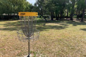 A disc golf basket stands in a grassy area surrounded by trees, under a clear sky.