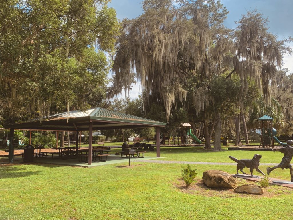 A park scene featuring a shaded picnic area with a green roof, surrounded by trees and grass, with a playground visible in the background and a dog statue in the foreground.