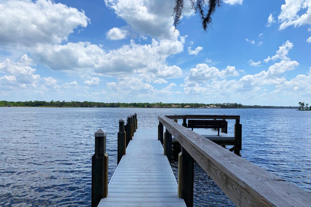 A wooden dock extends into a calm body of water under a bright blue sky with fluffy white clouds.