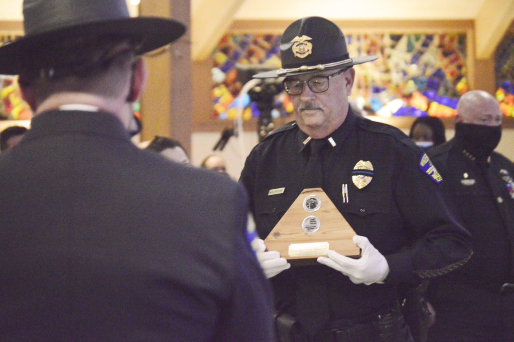 A police officer in uniform holds a wooden triangular plaque while facing another individual in a hat, surrounded by a group of people in a solemn setting.