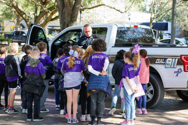 A group of children in purple shirts gathers around a police officer at a pickup truck, engaging in a discussion.