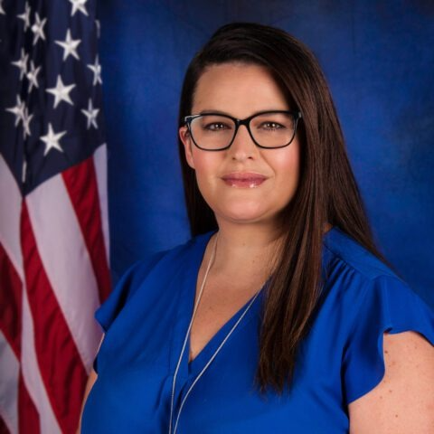 A woman with long dark hair and glasses, wearing a blue blouse, stands in front of a backdrop featuring the American flag.