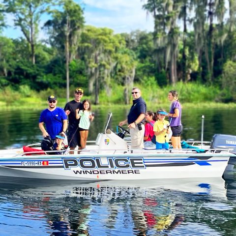 A group of people, including children and adults, are standing on a police boat labeled 'WINDERMERE' on a calm body of water surrounded by lush greenery.