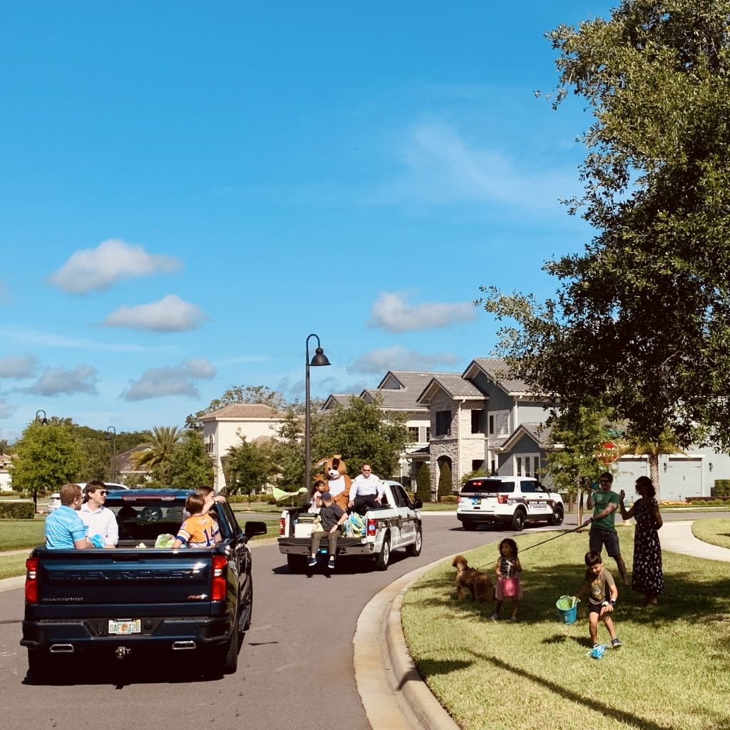 A neighborhood scene featuring several vehicles, including a pickup truck and a police car, with people gathered outside, some children playing on the grass, and houses in the background under a clear blue sky.