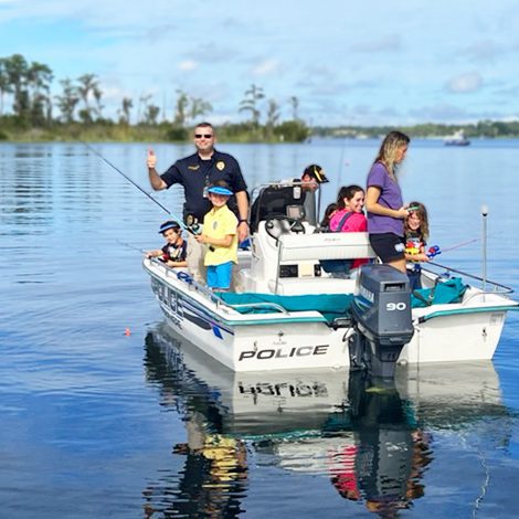 A police officer and several children are fishing from a small boat on a calm body of water, surrounded by trees and a clear blue sky.