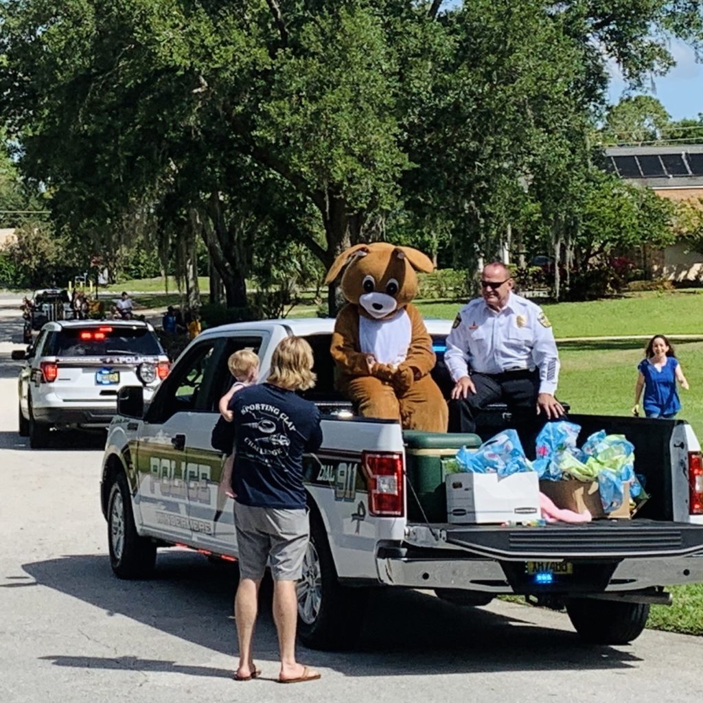 A child interacts with a person in a dog costume sitting in the back of a pickup truck, while an officer looks on, surrounded by a community event atmosphere.