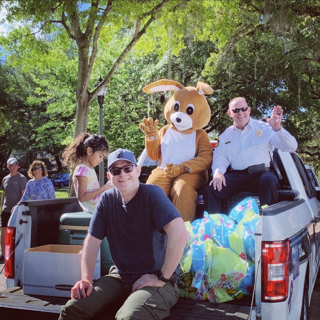 A group of people, including a person in a bunny costume, are sitting in the back of a pickup truck surrounded by colorful bags, with trees and a crowd in the background.