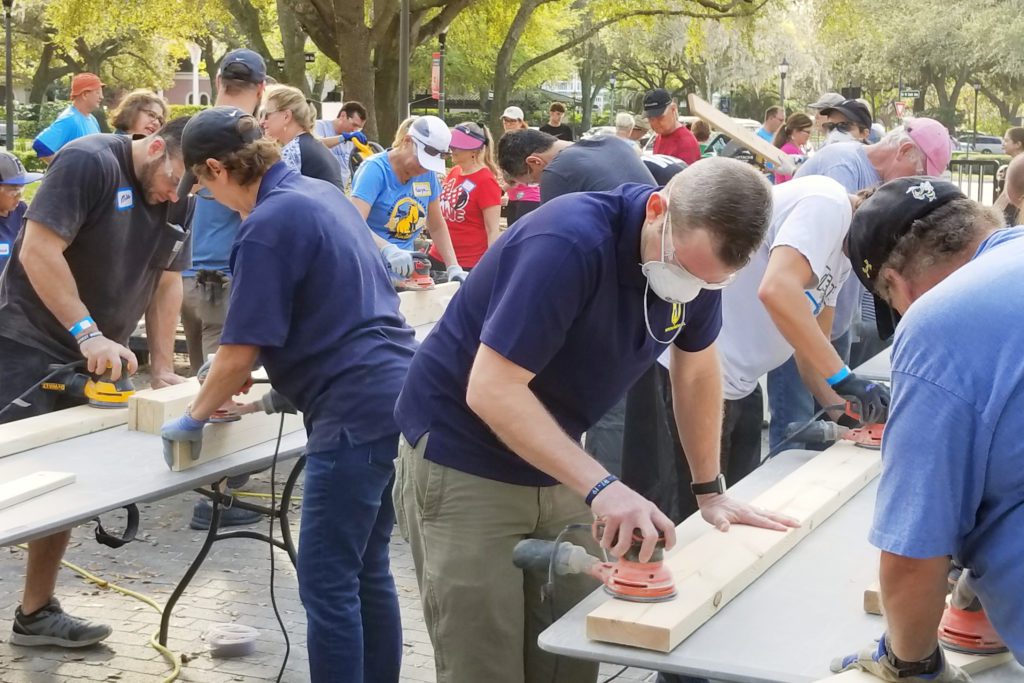 A group of people working together outdoors, using power sanders on wooden boards at a community event.