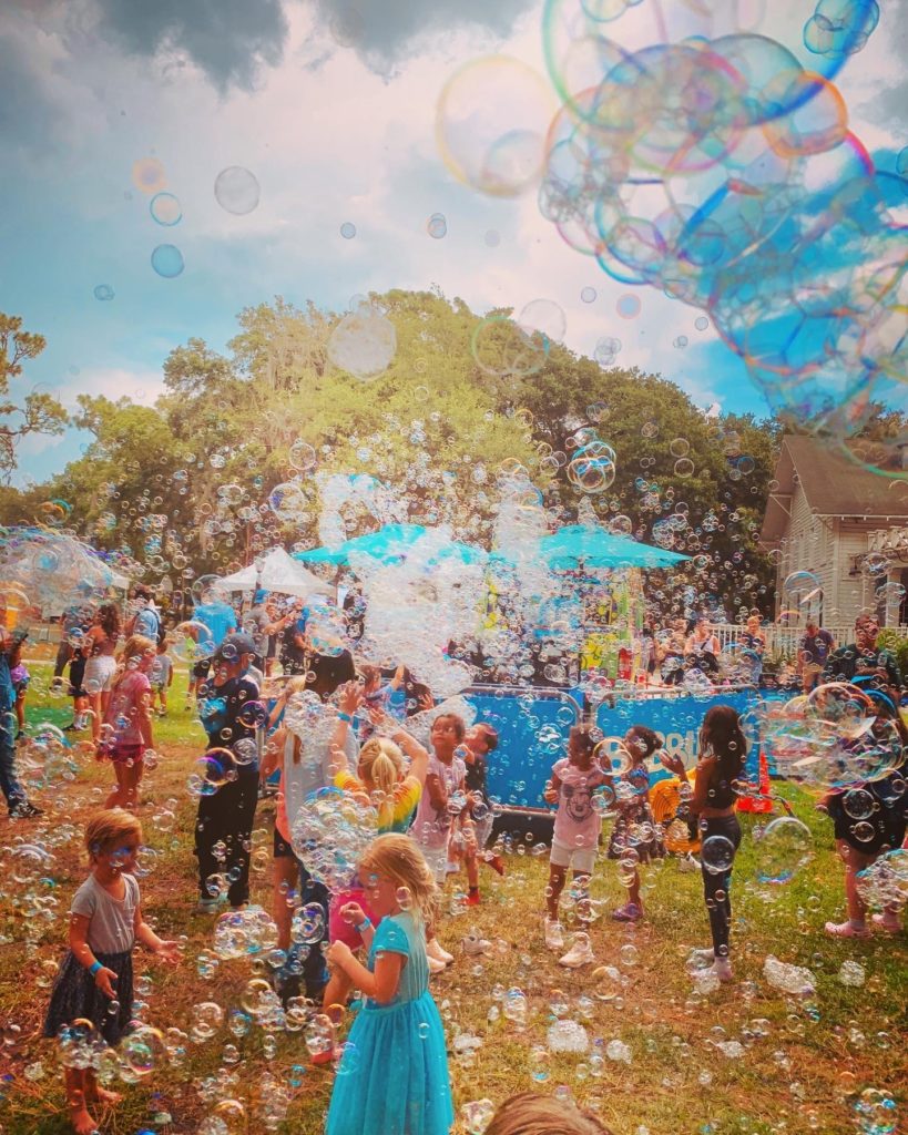 A lively outdoor scene filled with children playing among large bubbles, with a crowd enjoying a festive atmosphere under a partly cloudy sky.
