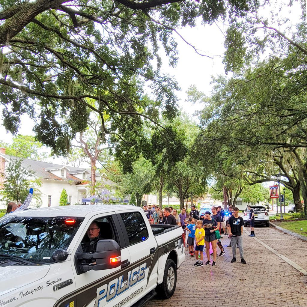 A police vehicle is parked on a tree-lined street, with a group of children gathered nearby, suggesting a community event or interaction with law enforcement.