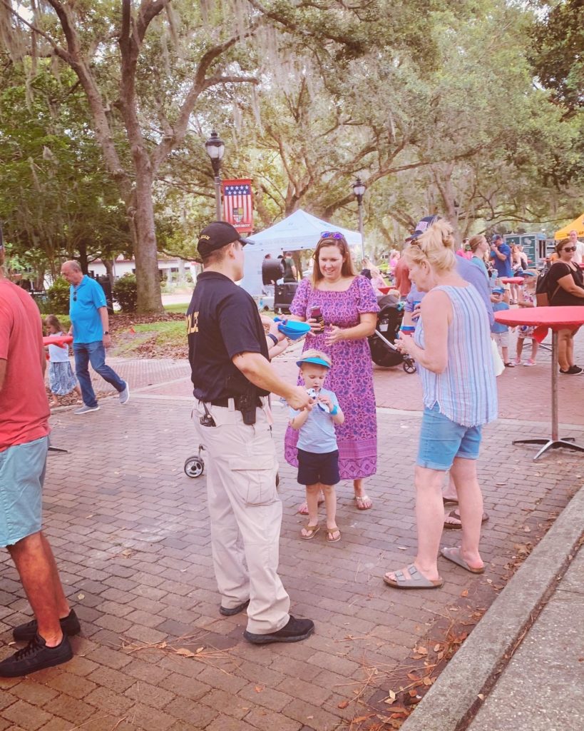 A police officer interacts with a woman and a child in a lively outdoor setting, surrounded by people enjoying a community event.