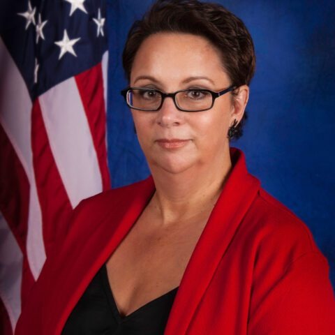 A woman wearing glasses and a red blazer poses in front of an American flag, with a serious expression. 