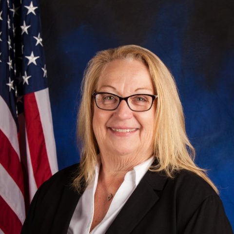 A woman with blonde hair wearing glasses and a black blazer poses in front of an American flag, smiling. 